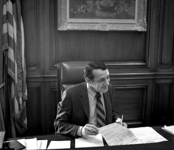 Black and white photo of a man in a suit working at a desk in an ornate government office with an American flag visible