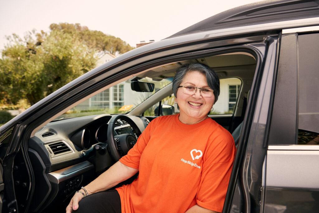 Driver wearing orange HopSkipDrive t-shirt sitting in driver's seat of vehicle, smiling at camera
