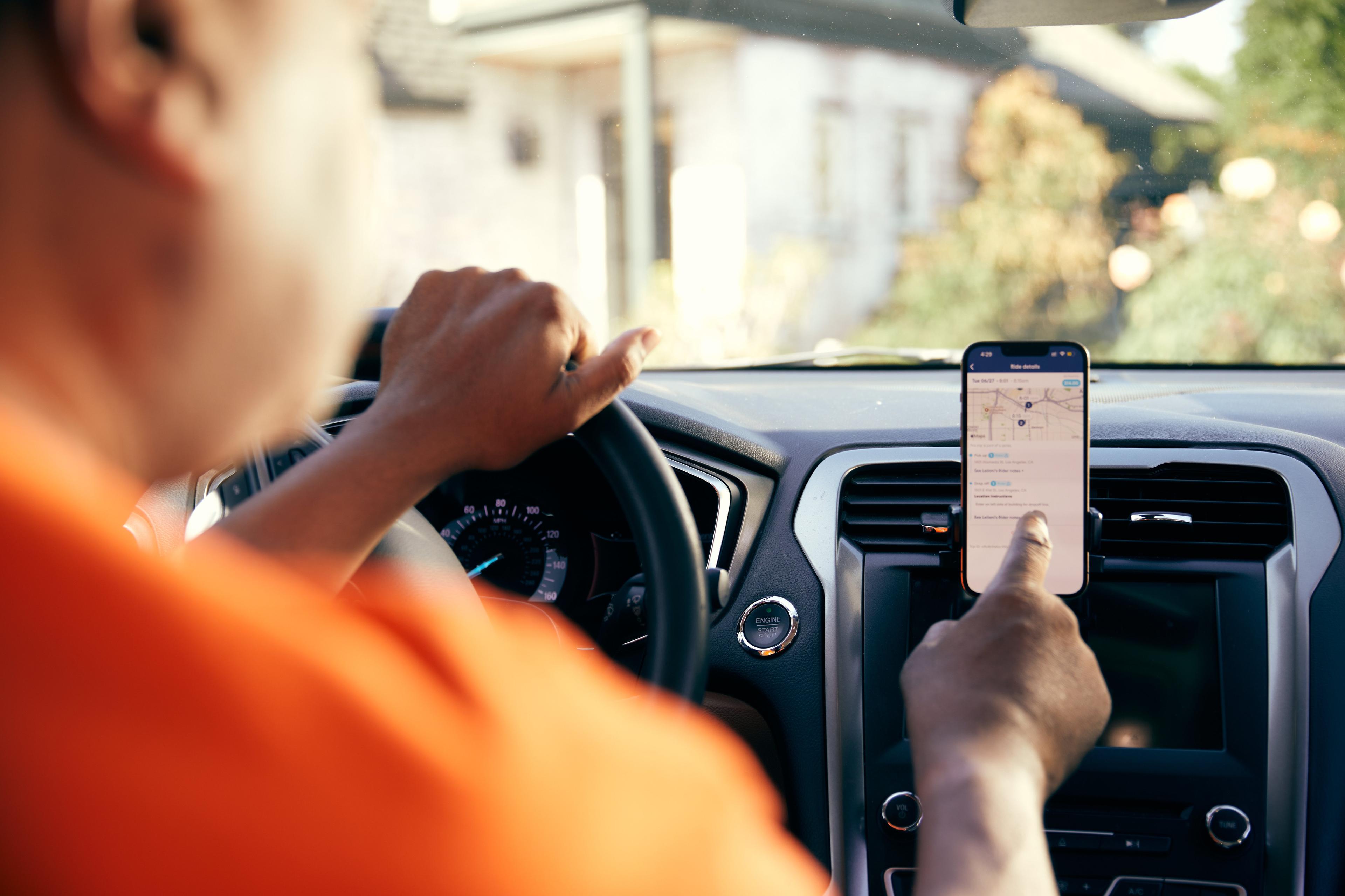 Driver in orange shirt checking navigation app on smartphone mounted to car dashboard while holding steering wheel