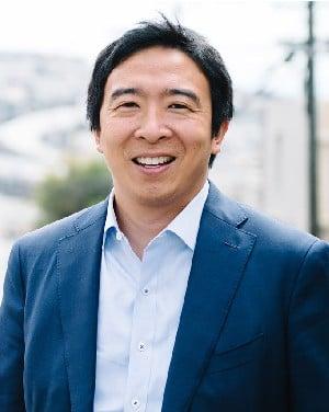 Professional portrait of a person in a navy blue suit and white shirt, smiling warmly against a soft-focused outdoor background