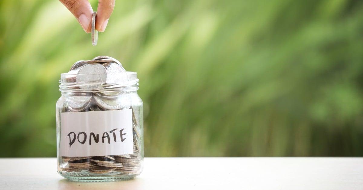 Hand dropping coin into glass jar labeled 'DONATE' filled with silver coins, against blurred green background