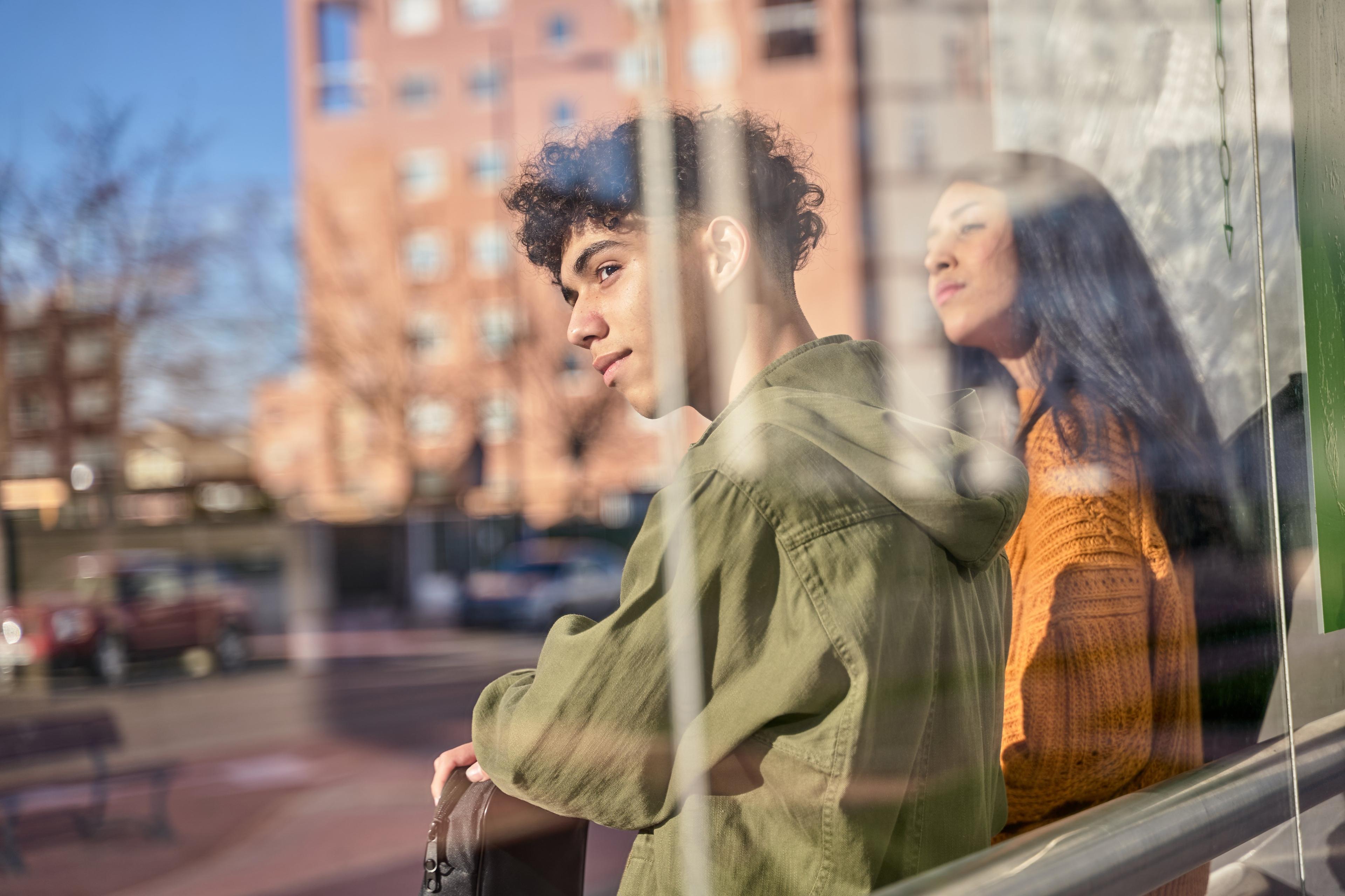 Two young people looking through window, one in green jacket, one in orange sweater, with city buildings reflected in glass
