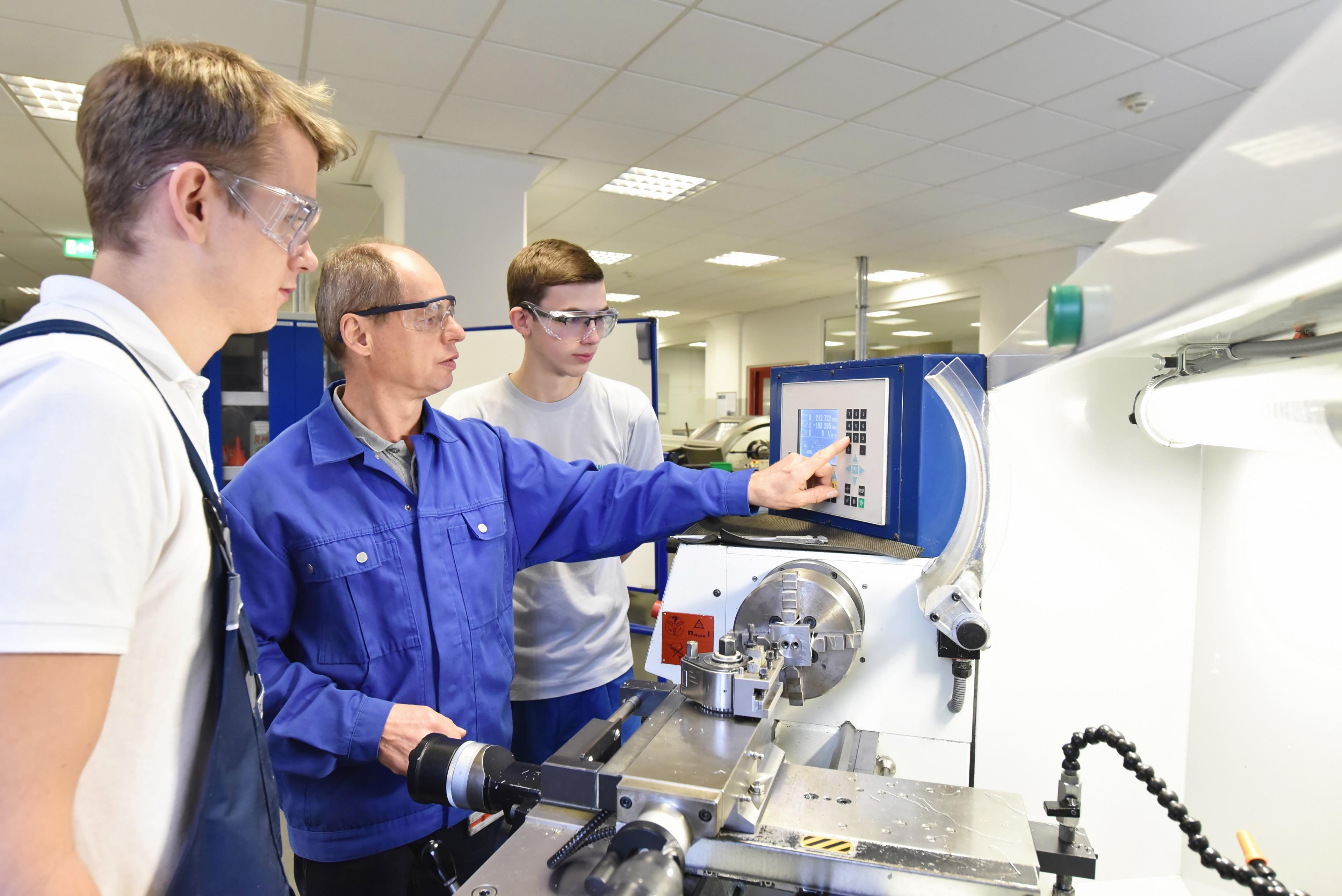 Instructor in blue coveralls demonstrating CNC machine operation to two students wearing safety glasses in a technical training facility
