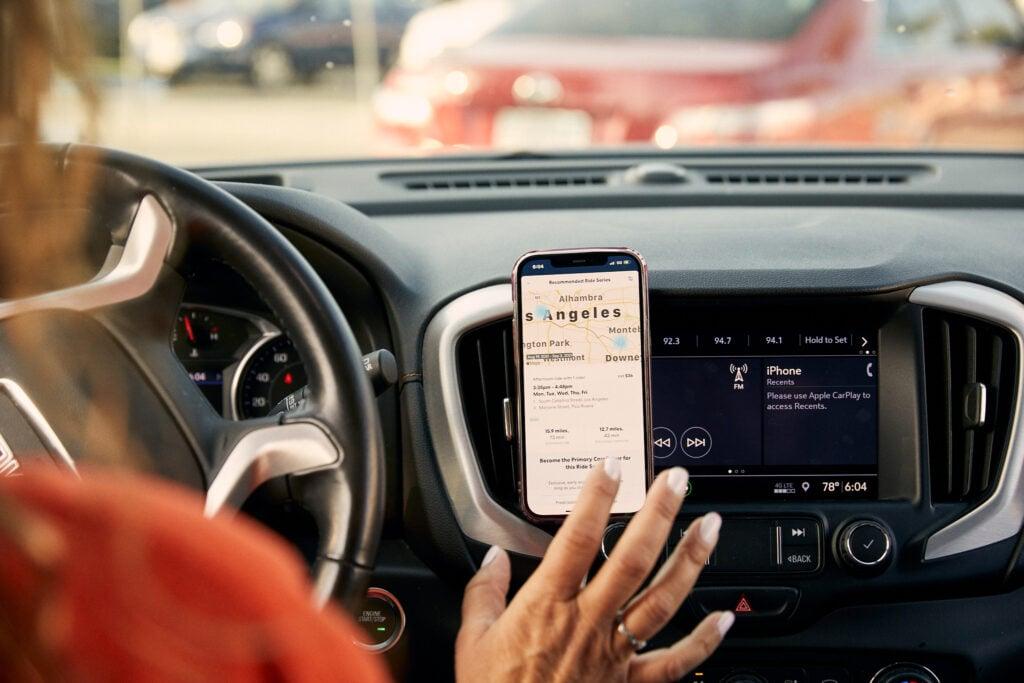 Hand touching smartphone mounted on car dashboard showing navigation map to Los Angeles, next to vehicle's infotainment system