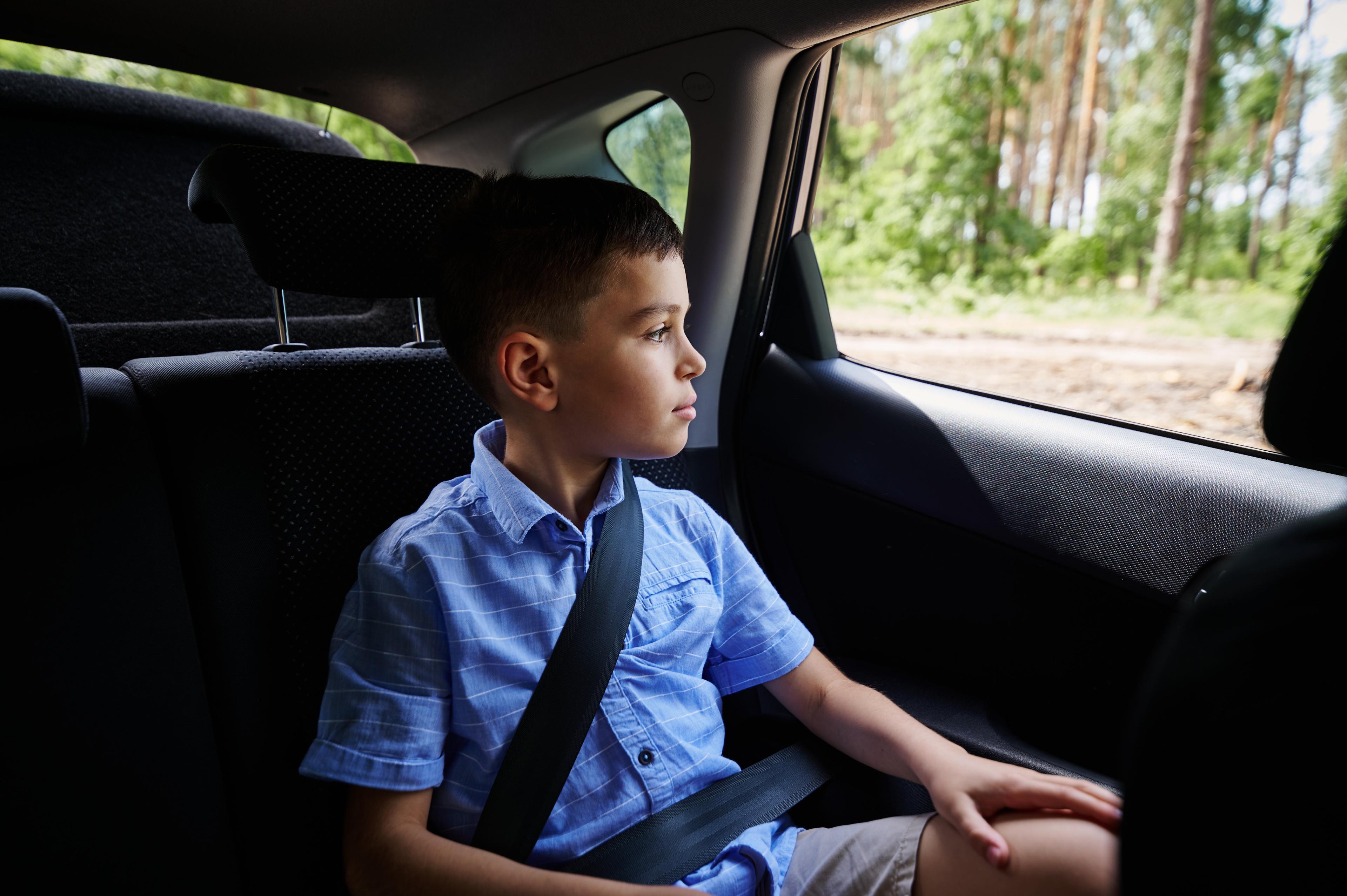 Young boy in blue shirt sitting in back seat of car, looking thoughtfully out the window while wearing seatbelt