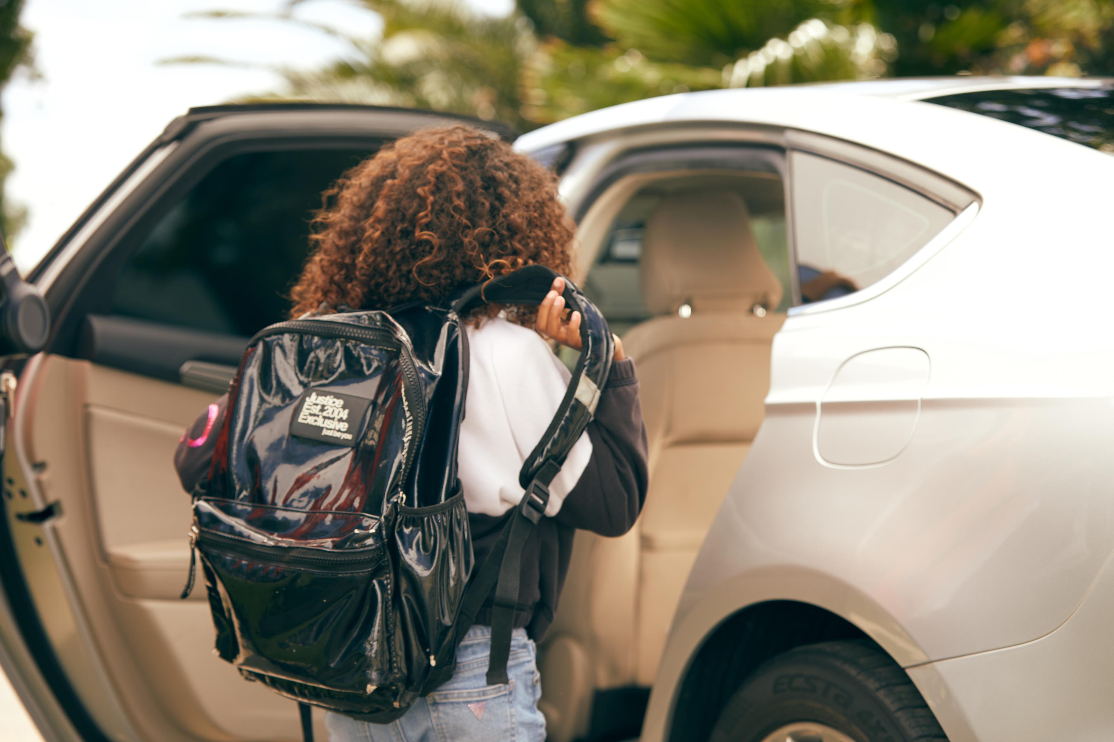 Person with curly brown hair wearing a black backpack getting into a white car with beige interior