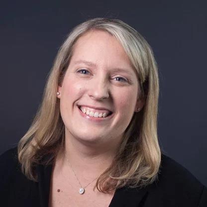 Professional headshot of a woman with blonde hair wearing a black blazer, smiling warmly against a dark background