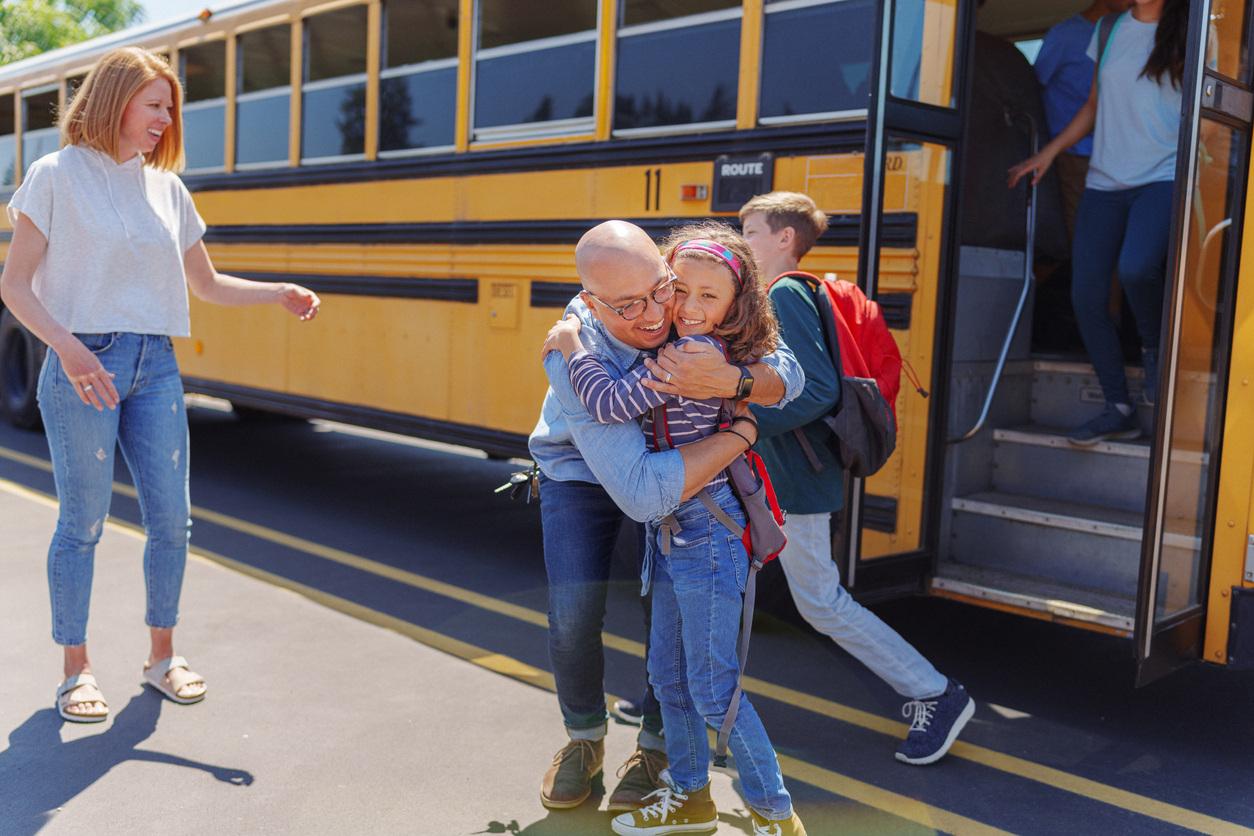 Heartwarming reunion scene at a school bus, with adult embracing student while others look on in the background