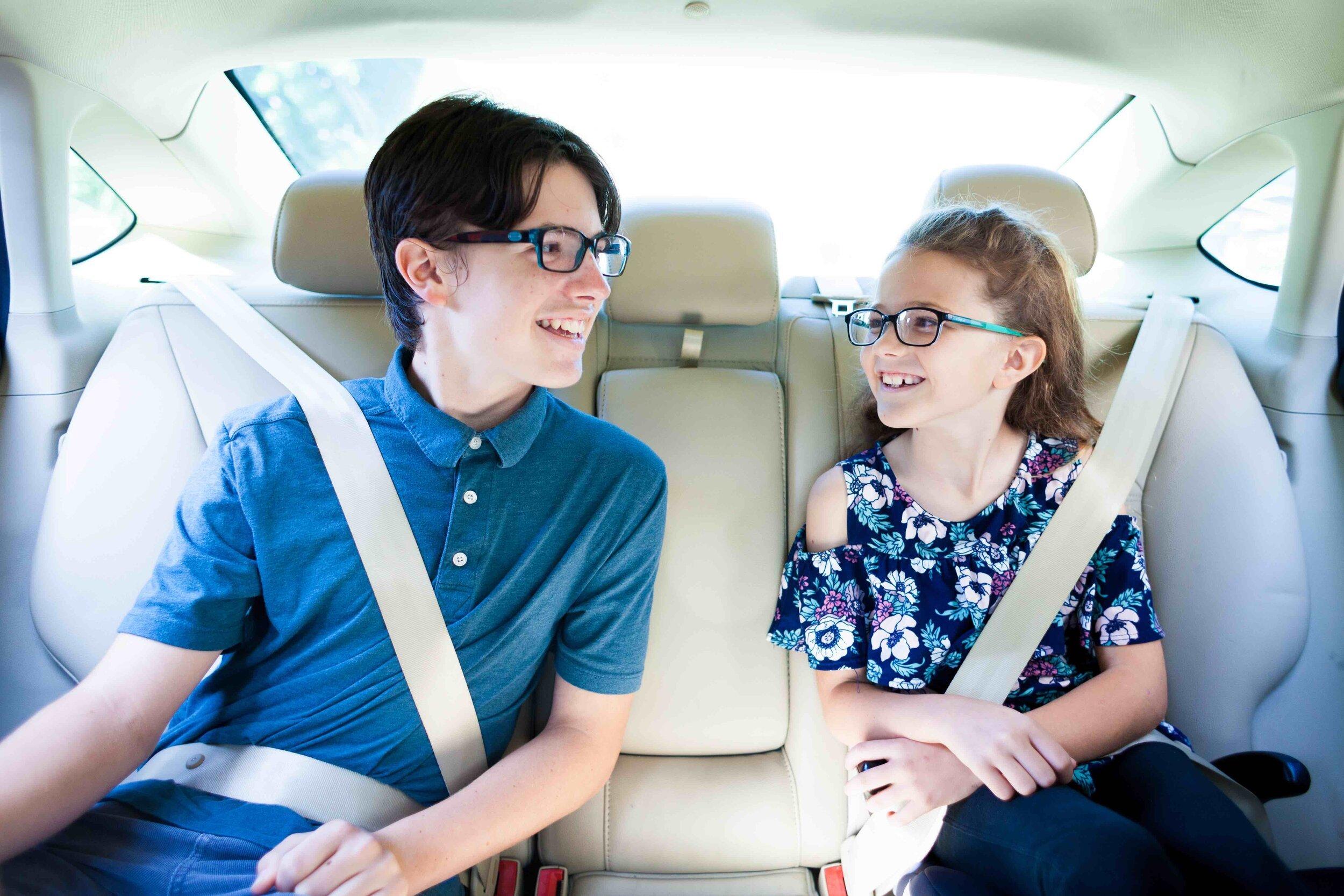 Two young siblings wearing glasses smile at each other while sitting in the backseat of a car with seatbelts fastened