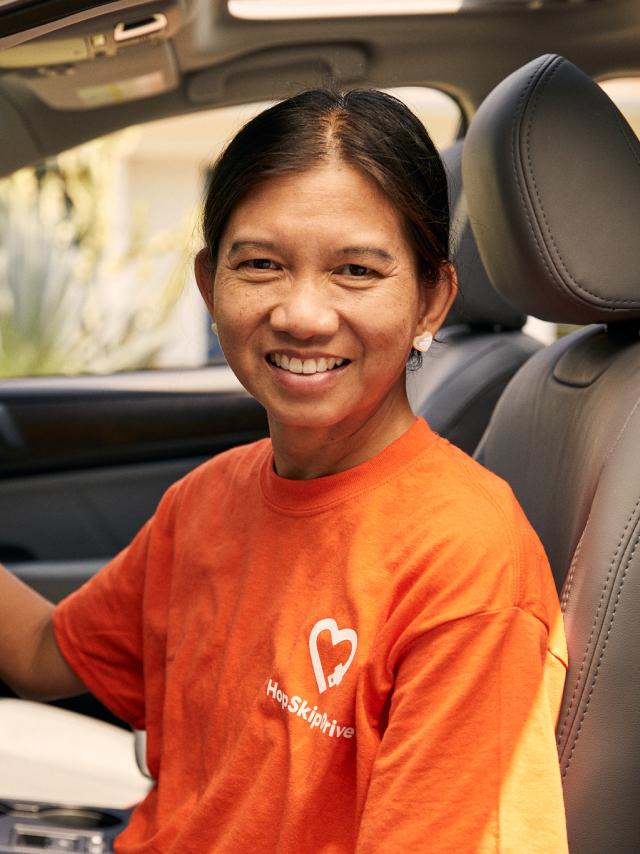 A smiling driver wearing an orange HopSkipDrive t-shirt seated in the driver's seat of a car