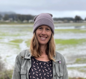Person wearing beanie and jacket smiling warmly against backdrop of natural wetland or tidal landscape