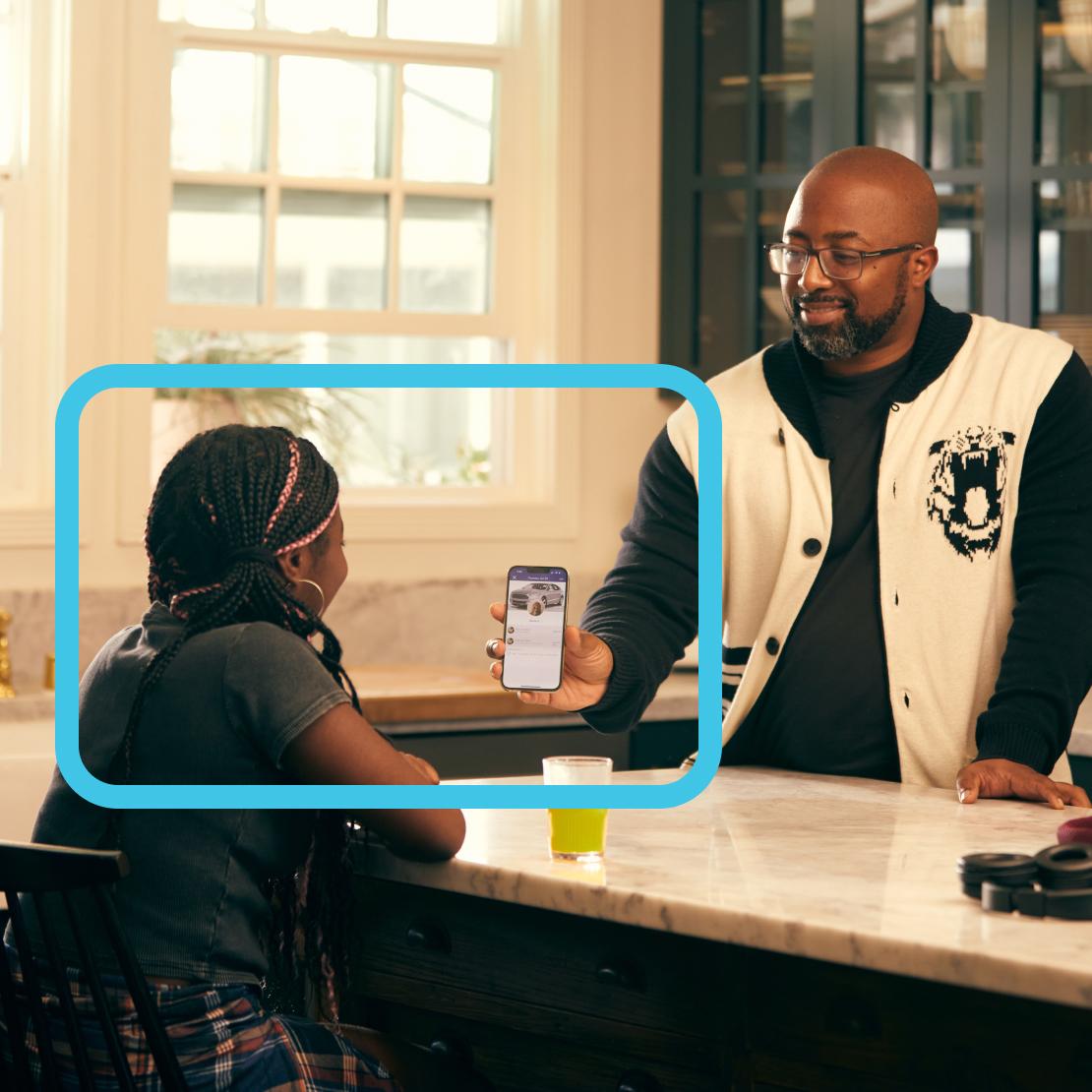 Two people having a conversation at a marble countertop while looking at a smartphone screen showing what appears to be a car image