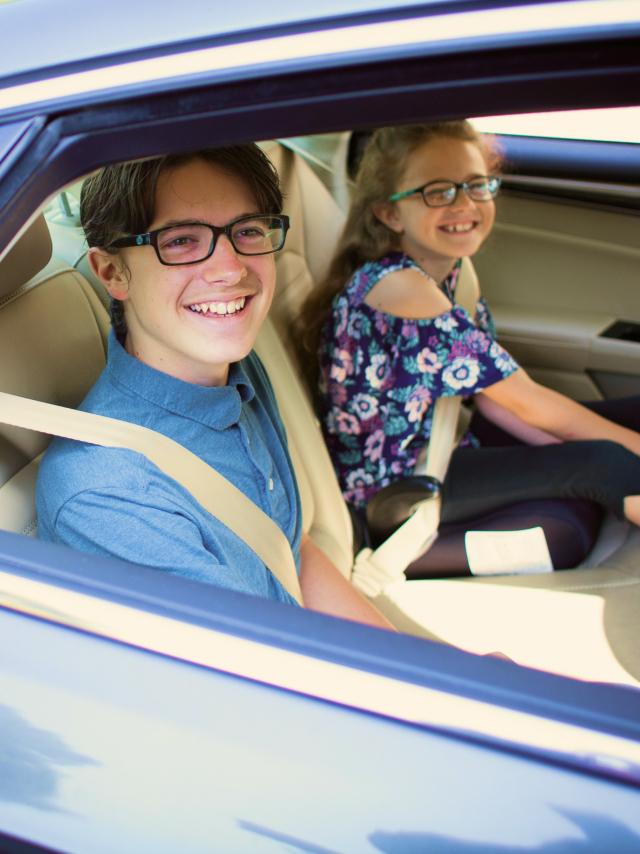 Two teenagers sitting in a car's front seats, both wearing glasses and seatbelts, smiling brightly