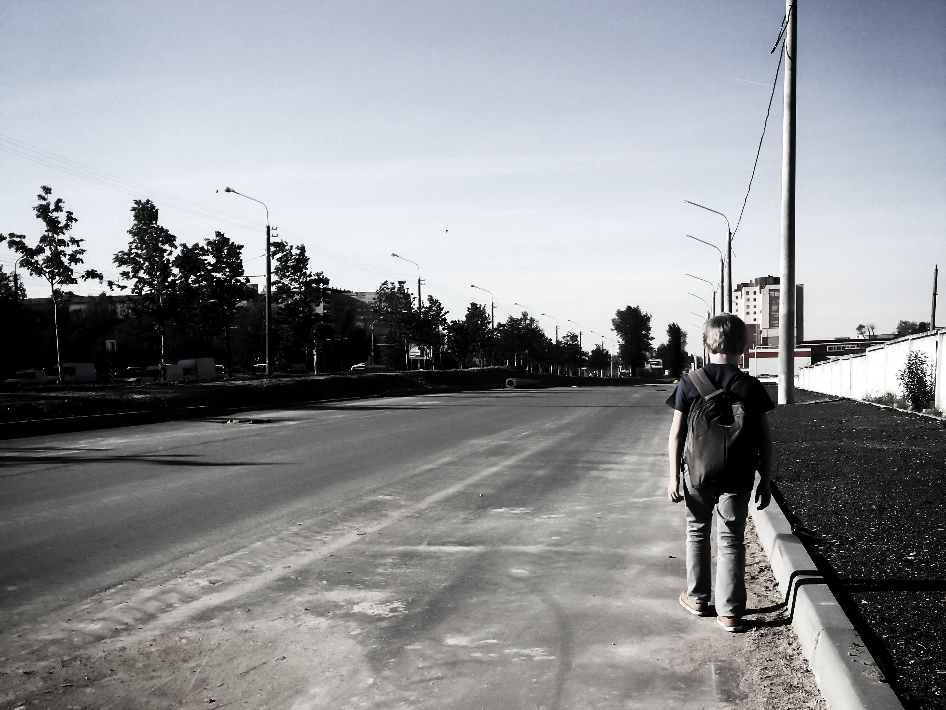 Person with backpack standing alone on empty street with street lights and trees, shot in black and white