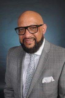 Professional in grey suit and patterned tie wearing black-framed glasses, smiling warmly against blue backdrop