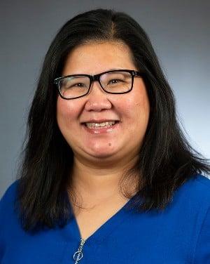 Professional headshot of a woman with glasses wearing a blue top, smiling warmly against a grey background