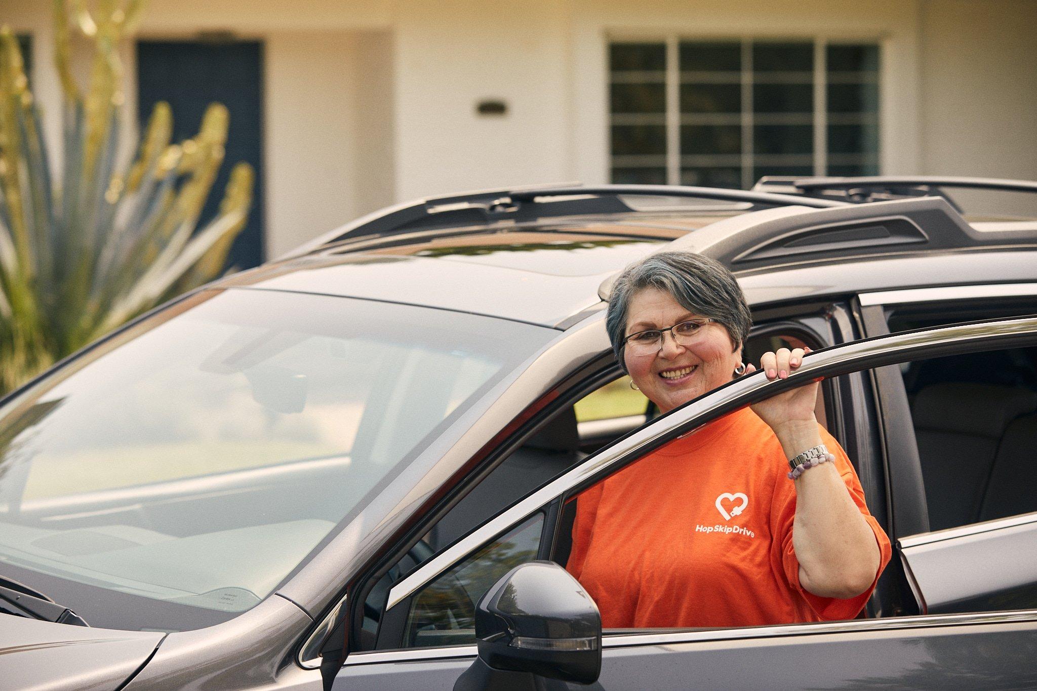 Driver wearing orange HopSkipDrive shirt smiling from driver's seat of silver SUV