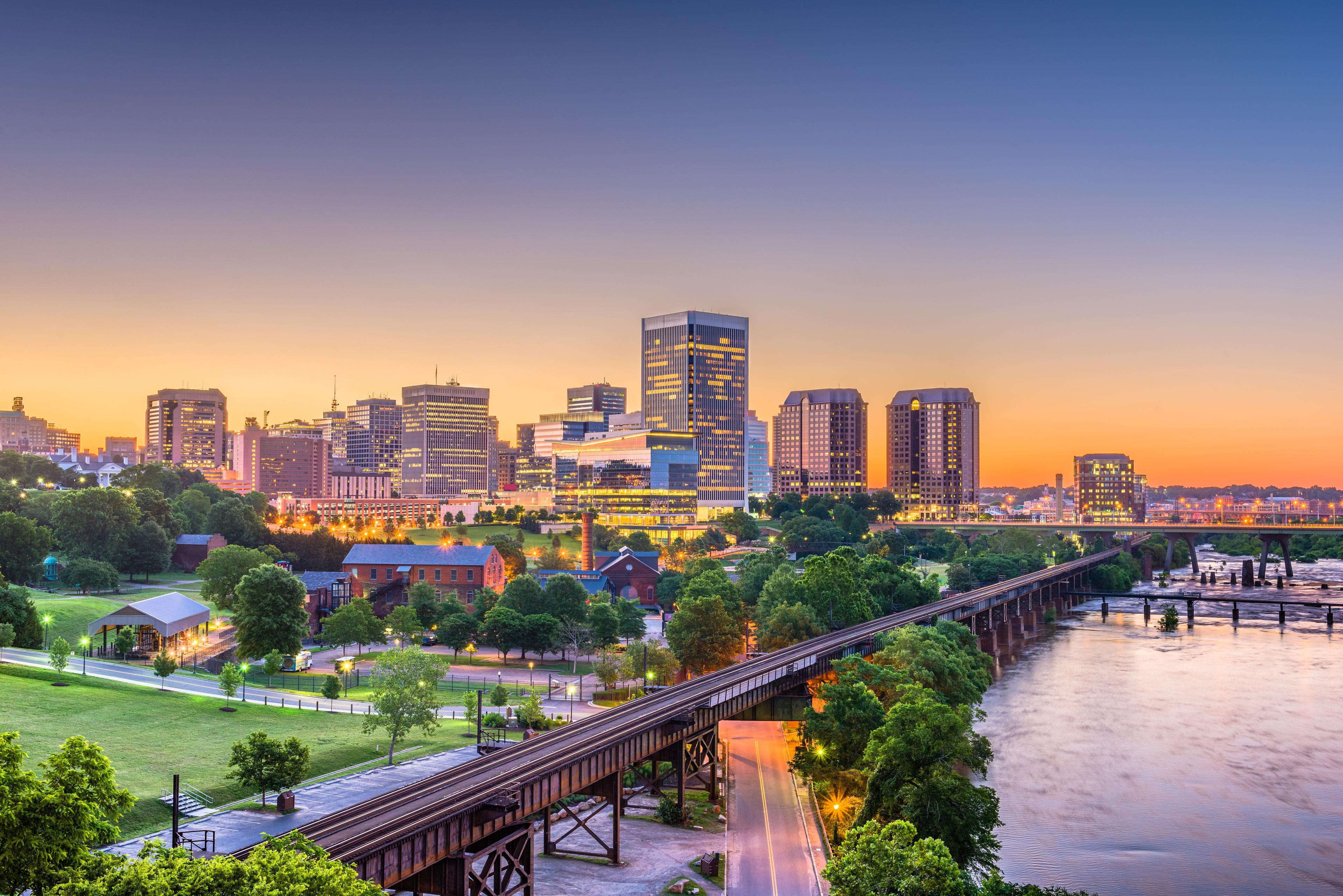 Richmond, Virginia cityscape at sunset with illuminated skyscrapers, railroad bridge, and James River surrounded by lush greenery