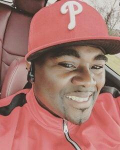 Close-up portrait of a person wearing a Philadelphia Phillies baseball cap and red jacket, smiling warmly at the camera