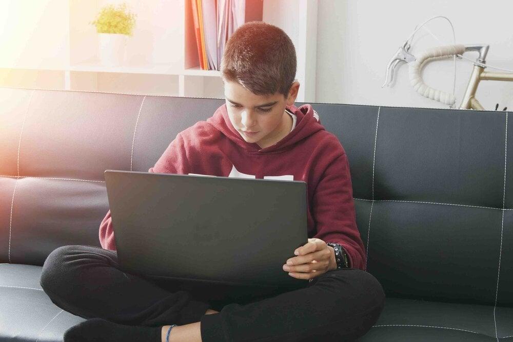 Young person in red hoodie sitting cross-legged on gray couch, focused on laptop screen