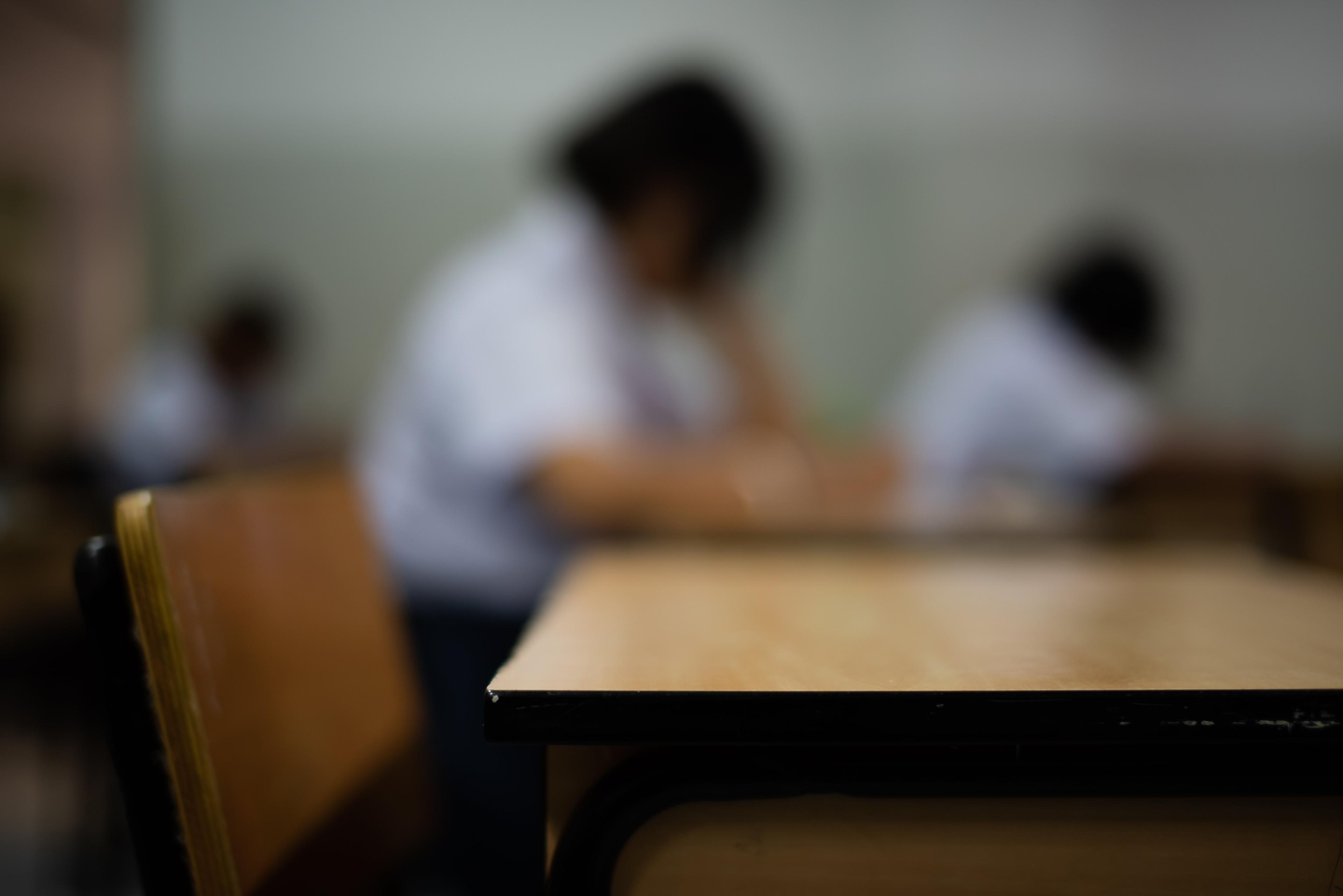Blurred view of students in white uniforms taking an exam, with empty desk and chair in sharp focus in the foreground