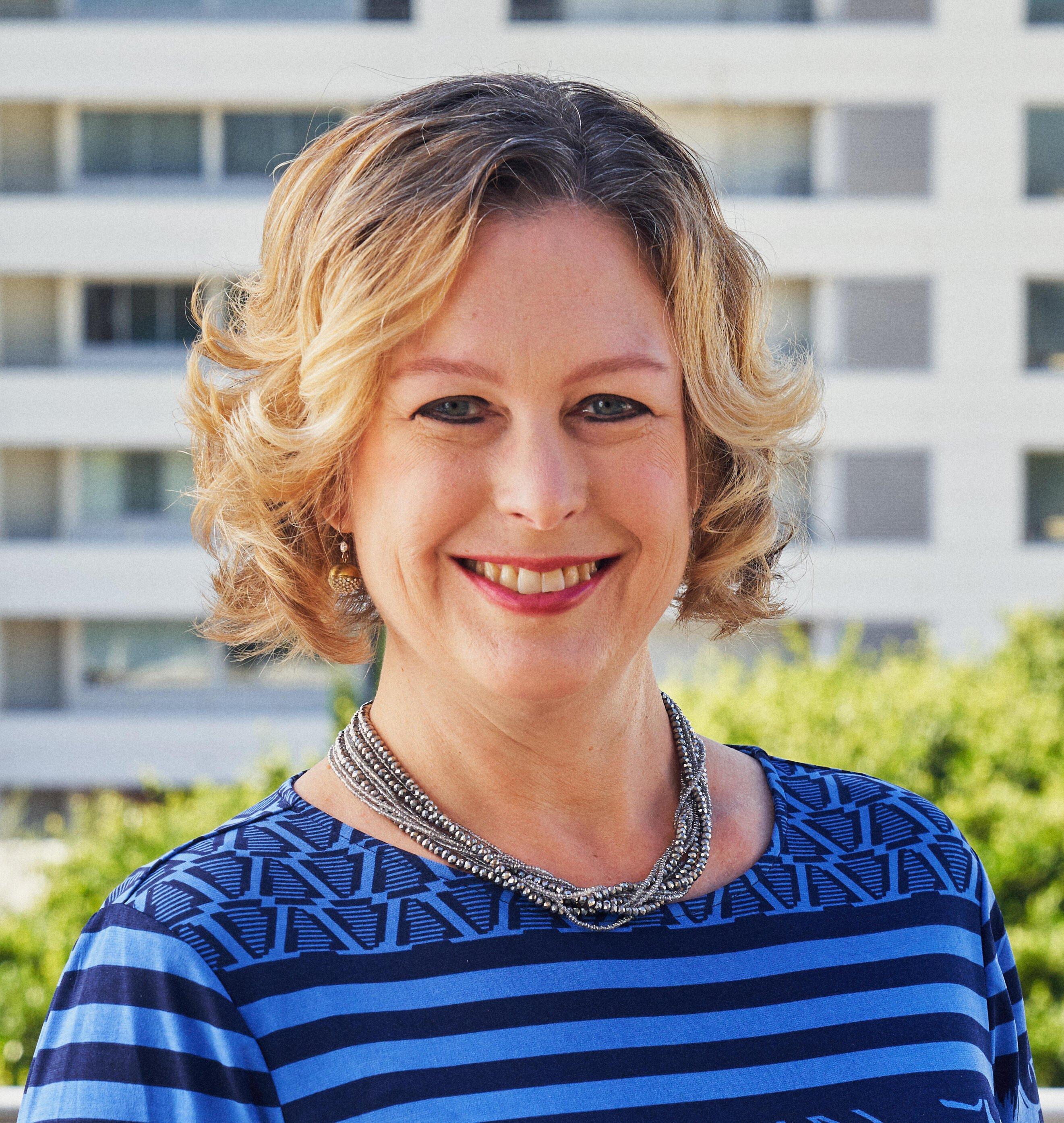 Professional portrait of a person wearing a blue striped top and silver necklace, smiling warmly against an office building background