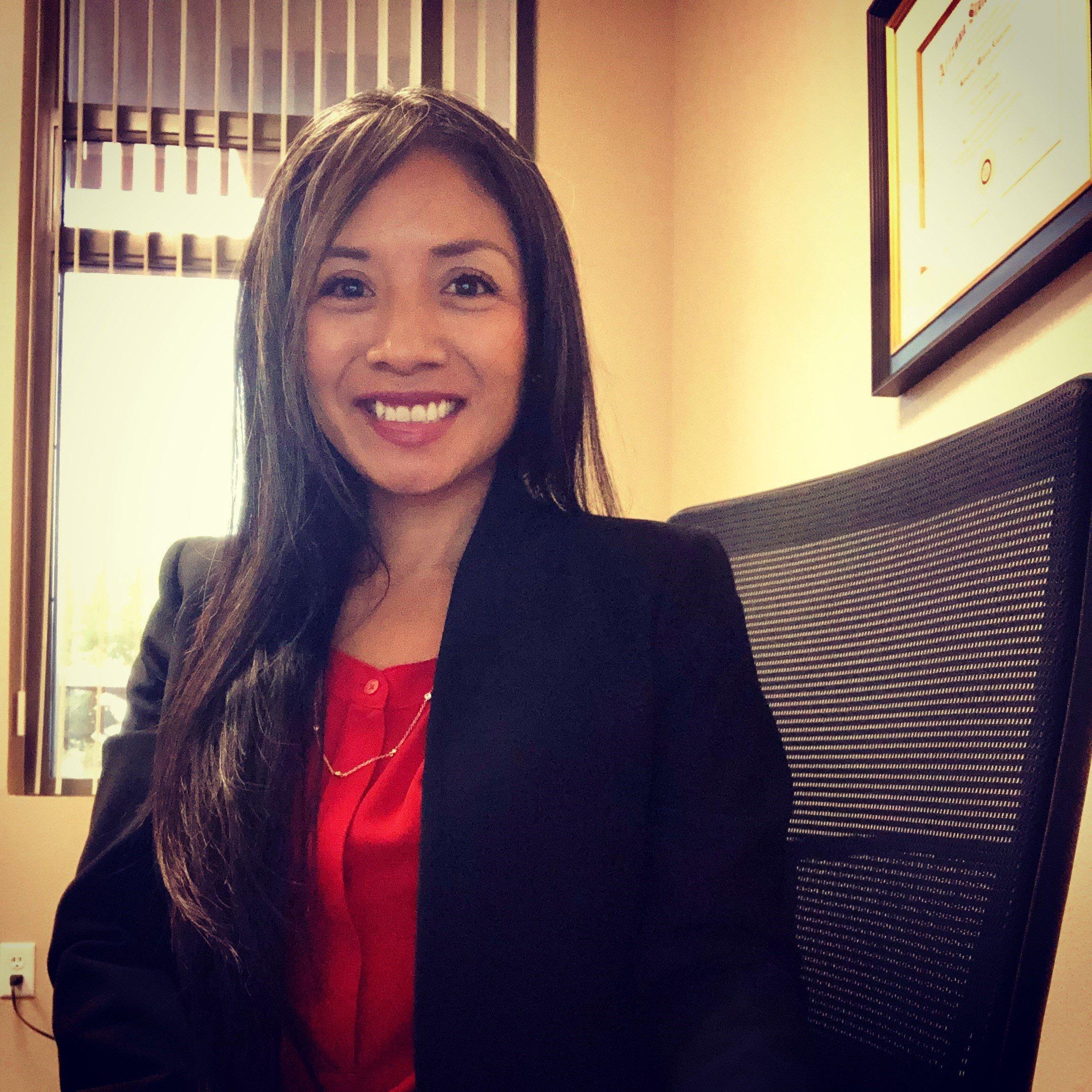 Professional woman wearing black blazer and red blouse smiling at camera in office setting with framed certificate visible