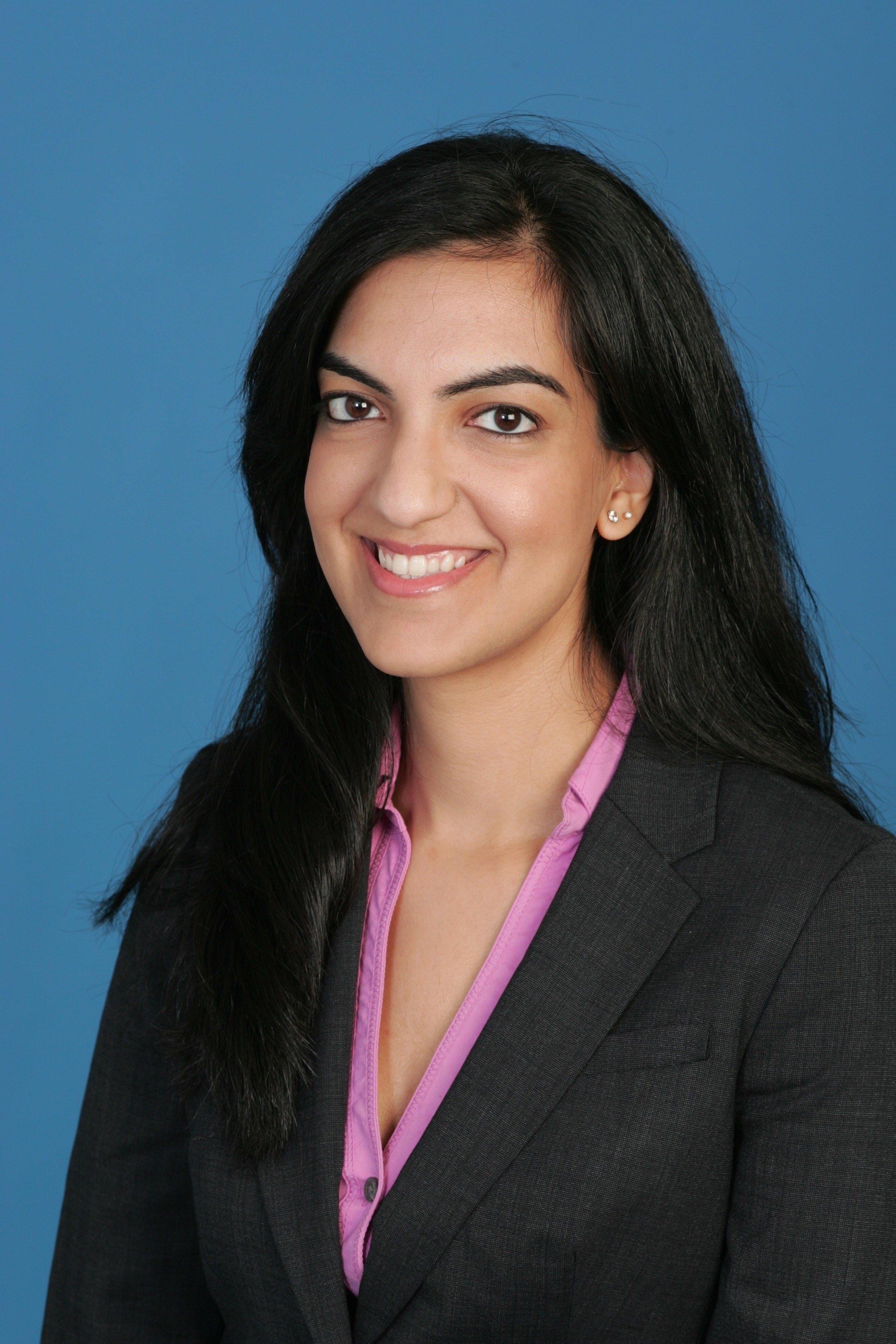 Professional portrait of a person in business attire with a dark blazer and pink shirt against a blue background