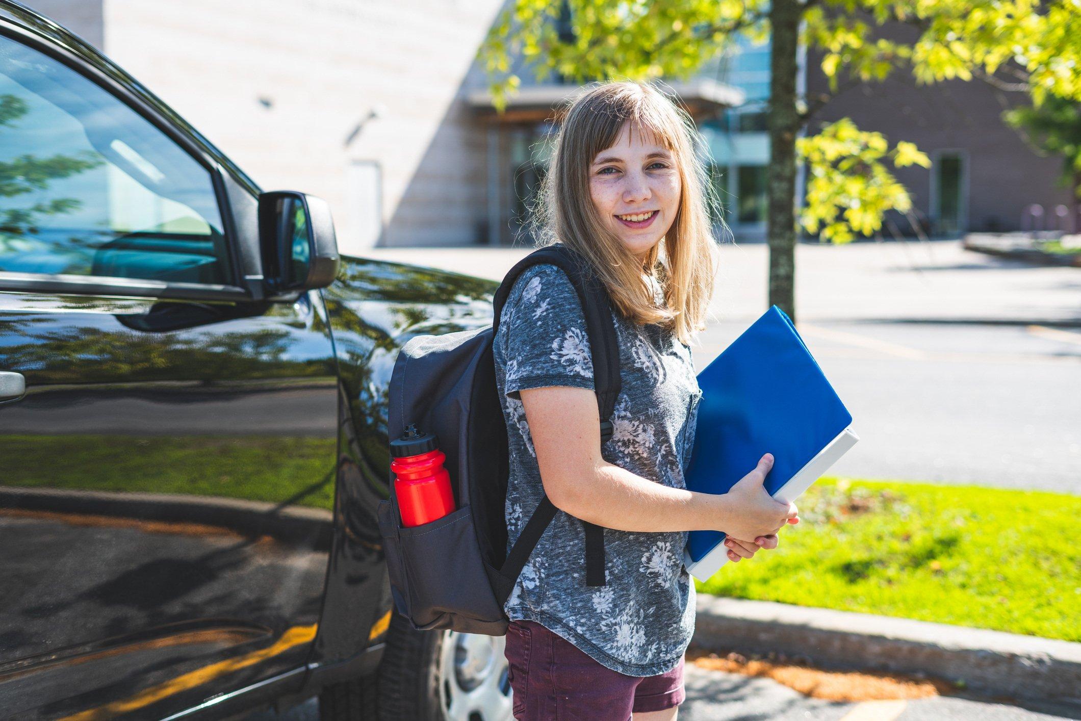Student with backpack and folder standing next to car on sunny campus, smiling at camera