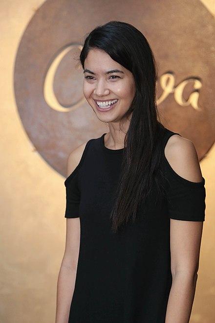 Professional portrait of a woman in a black cold-shoulder dress smiling warmly against a golden circular backdrop