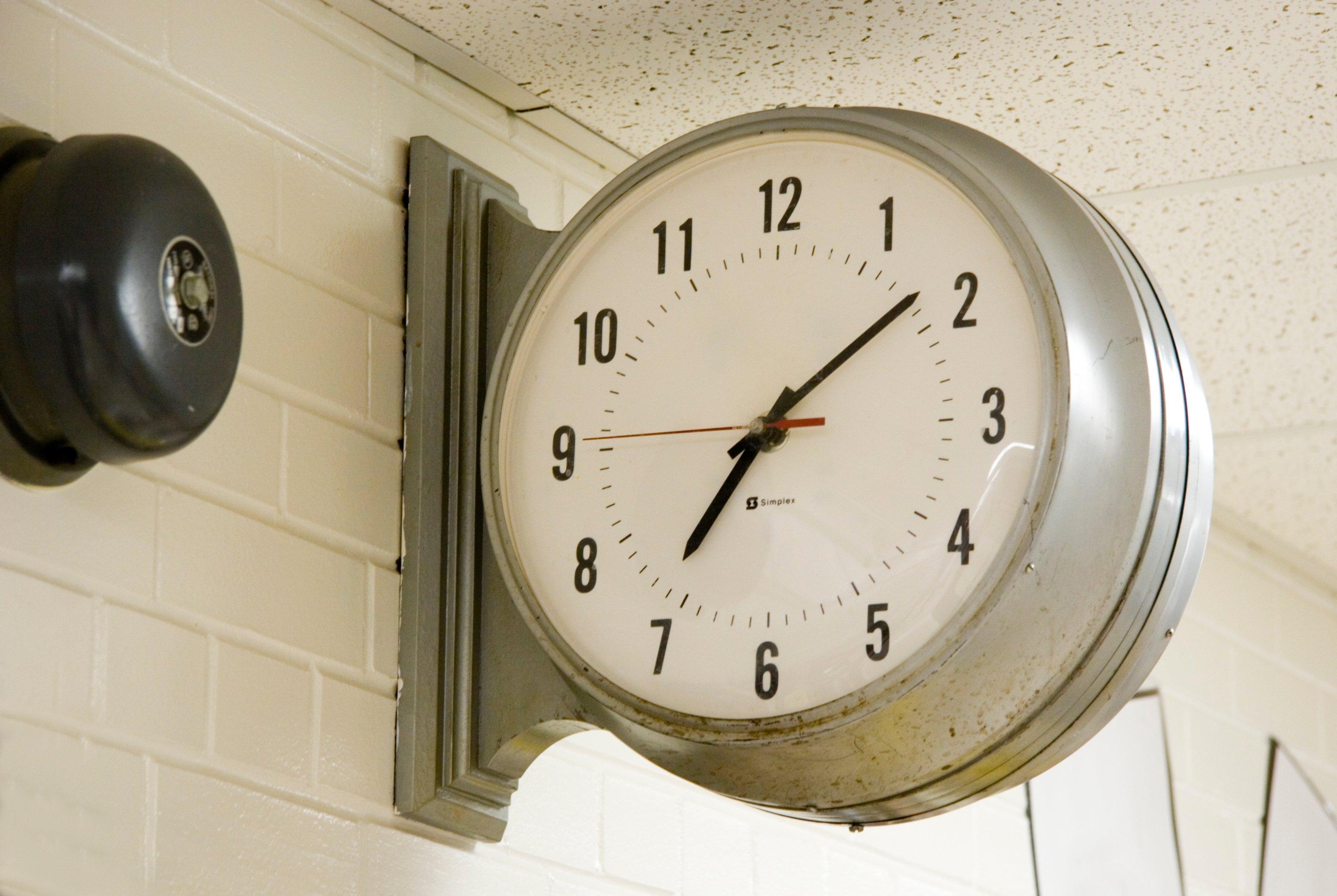 Double-sided institutional Simplex wall clock mounted on white brick wall showing approximately 5:35 time