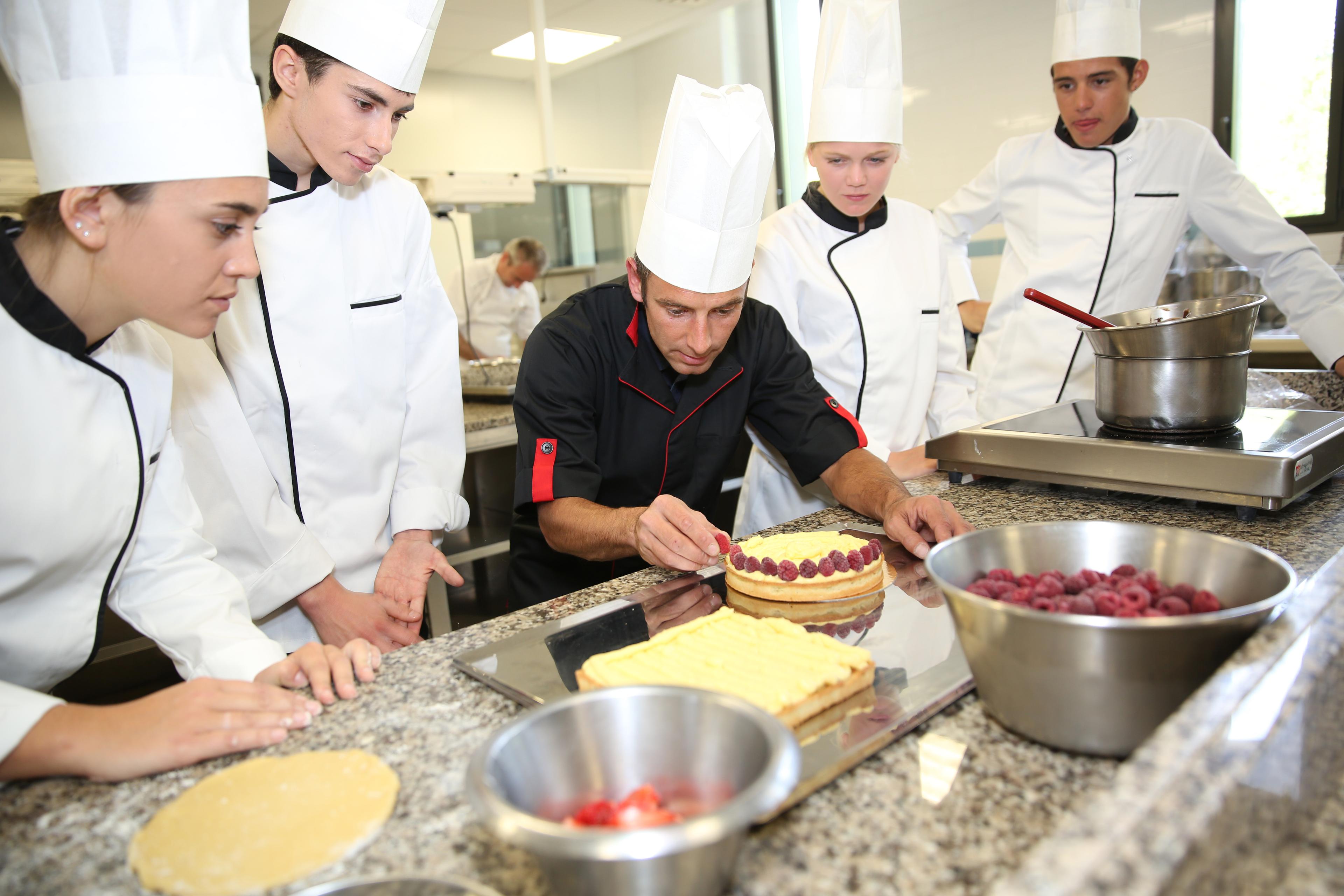 Chef in black uniform demonstrating cake decorating technique to culinary students in white uniforms and chef hats in professional kitchen