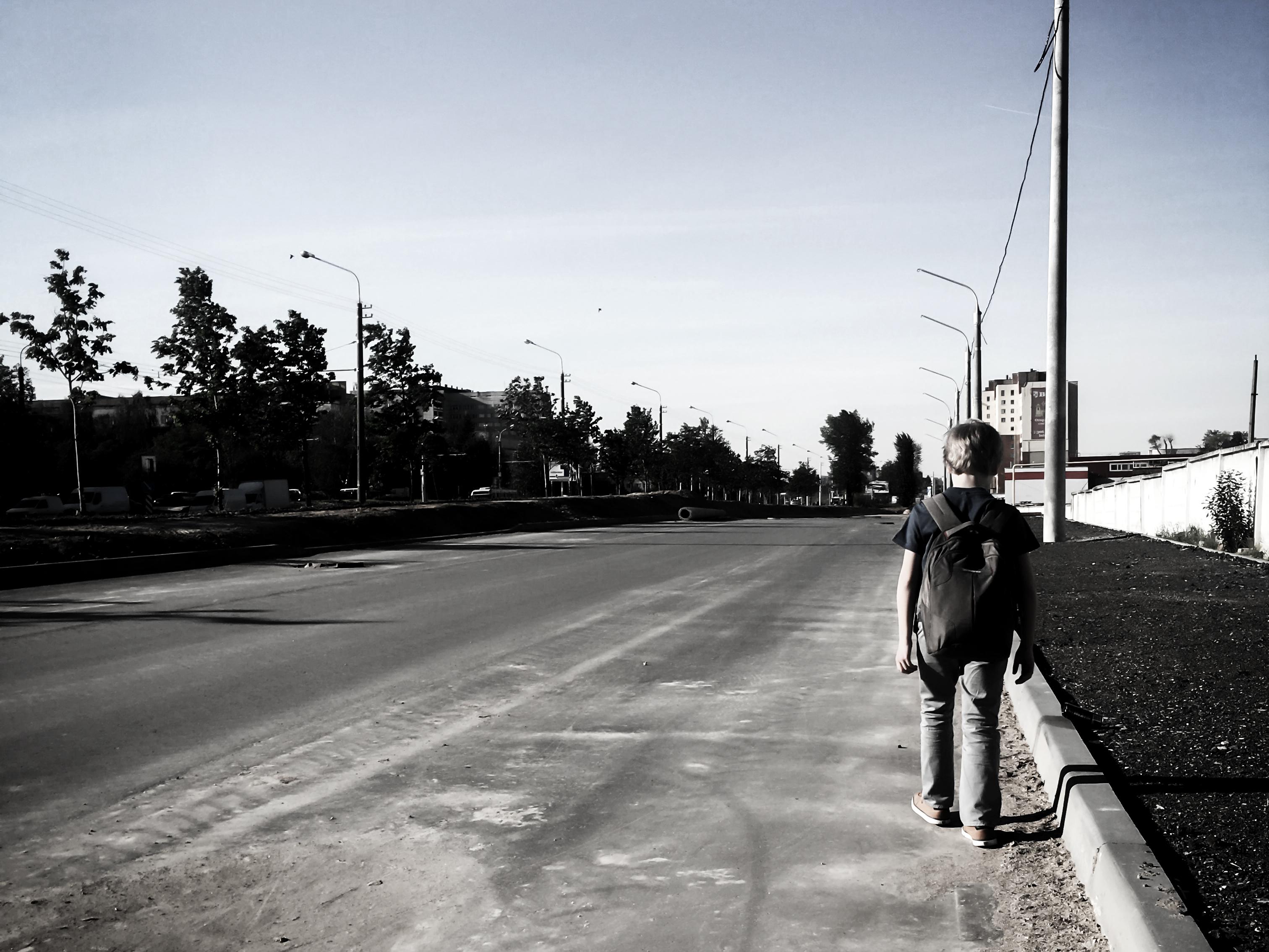 Person with backpack standing alone on the edge of an empty urban street lined with lamp posts and trees