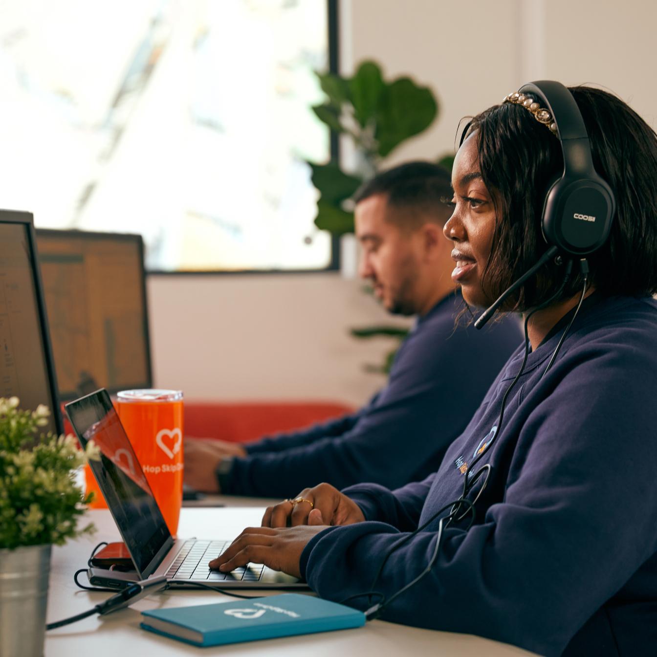 A person wearing headphones works on a laptop in a modern office space, with a colleague visible in the background and an orange drink cup on the desk