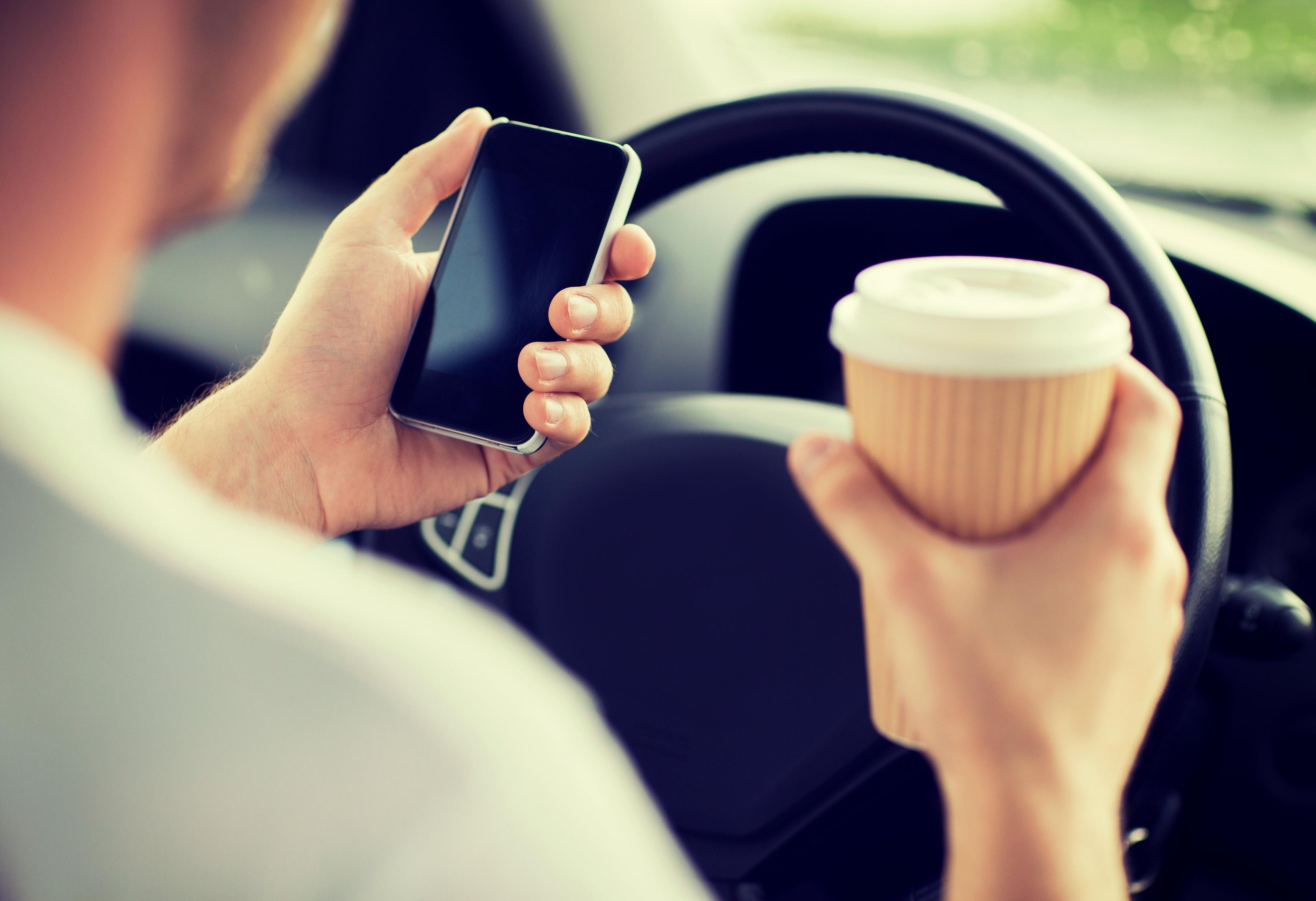 Driver holding smartphone and coffee cup while behind steering wheel, demonstrating dangerous distracted driving behavior