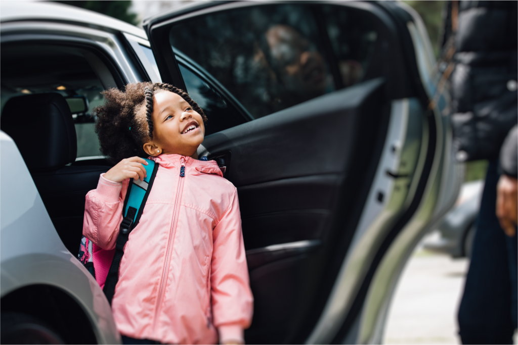 Child wearing pink jacket smiling brightly while getting out of car with backpack