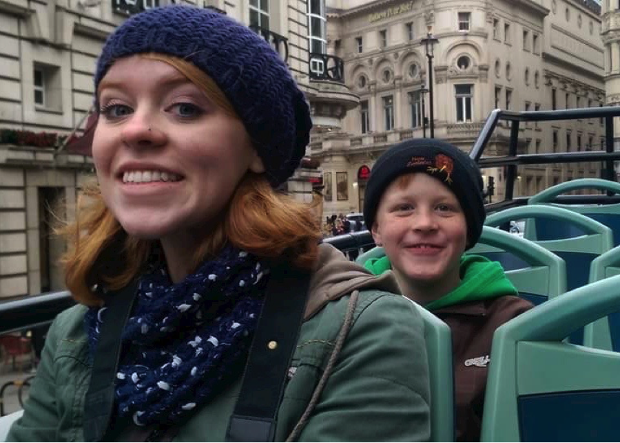 Two tourists wearing winter hats and coats smiling while sitting on upper deck of sightseeing bus with historic buildings in background