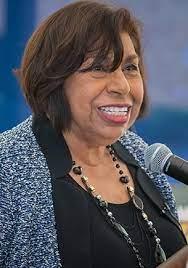 Woman in navy blazer and black necklace speaking into microphone with warm smile against blue background