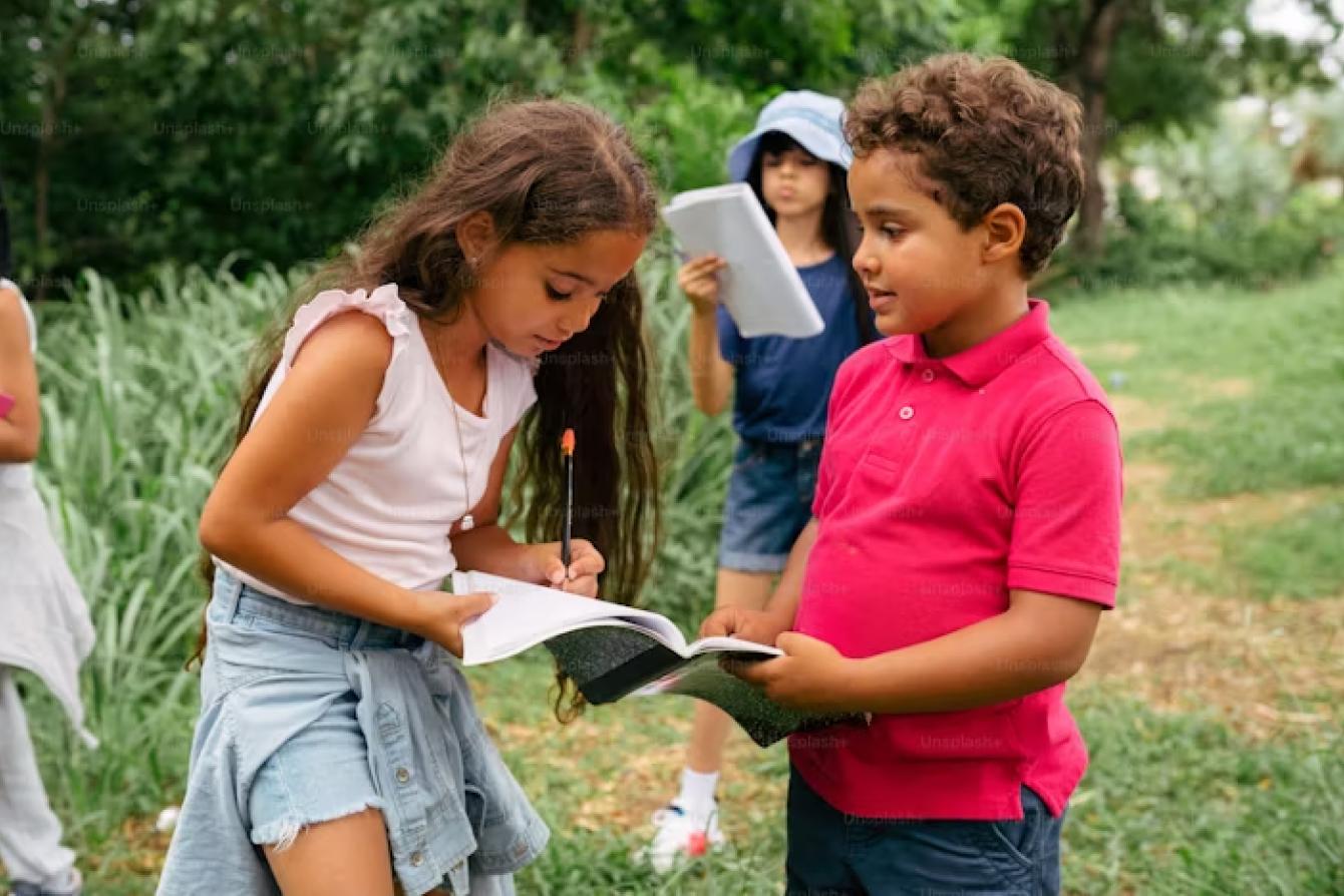 Two students collaborating on an assignment outdoors, with one writing in a notebook while another watches, and a third student visible in the background