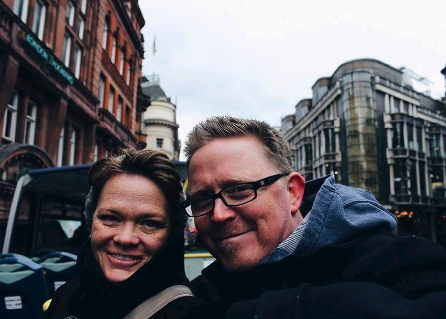 Close-up selfie of a couple smiling on a city street with Victorian architecture in the background