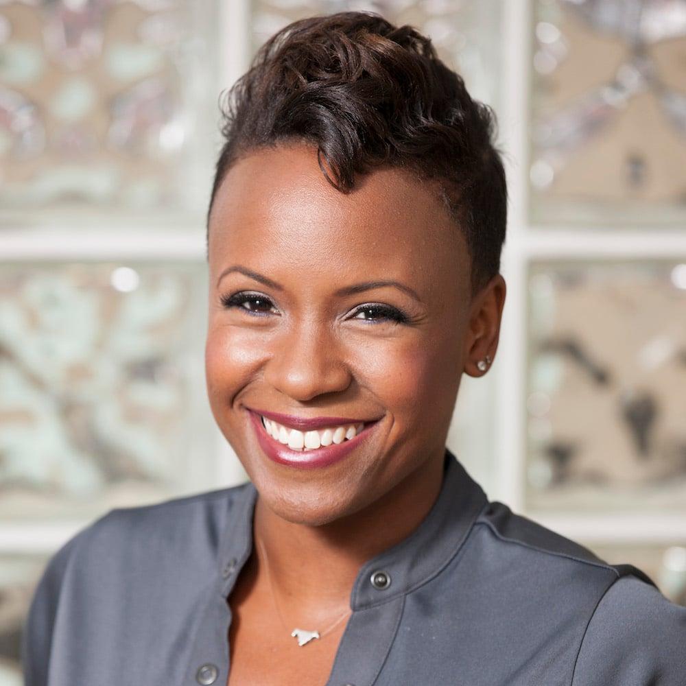 Professional headshot of a woman in a gray jacket, featuring a warm smile and short styled hair against a light patterned background