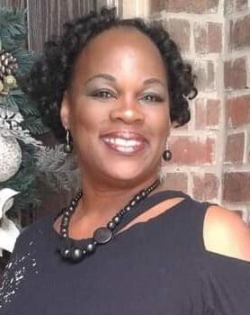 Professional portrait of a person in black attire with a beaming smile against a brick wall with holiday decorations