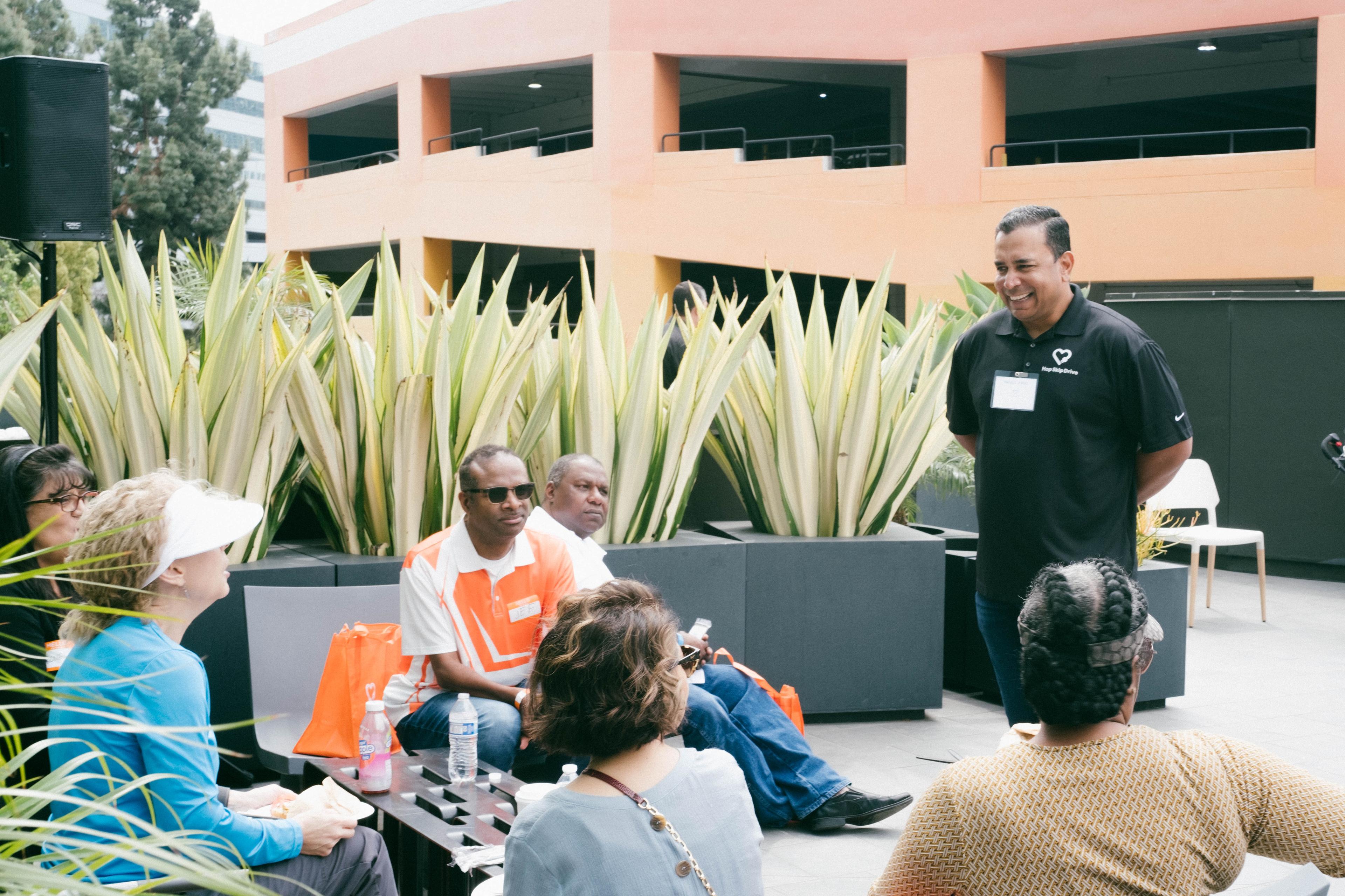 Speaker in black polo shirt addressing small group seated outdoors with decorative plants and parking structure in background