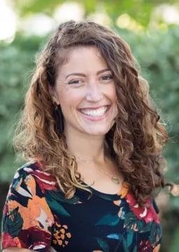 Professional portrait of woman with curly brown hair wearing floral print top, smiling warmly against blurred natural background