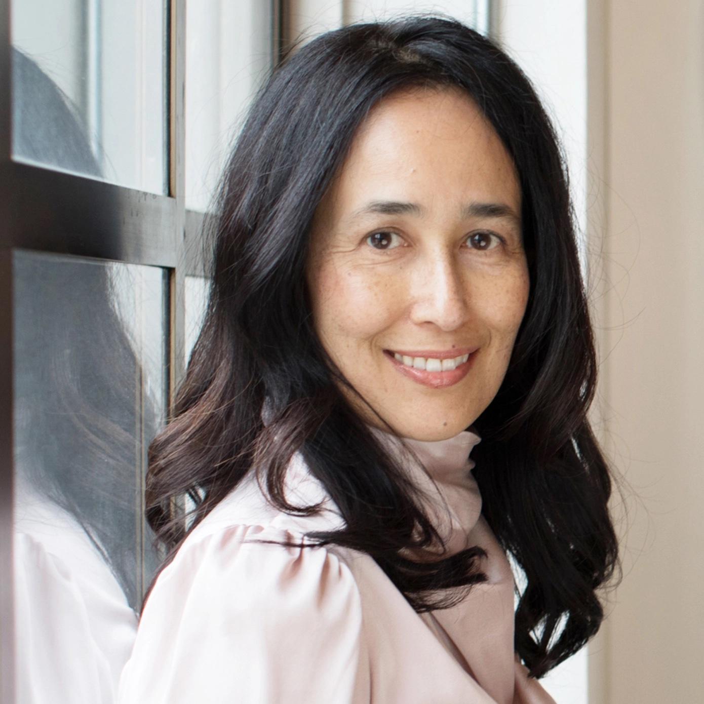 Professional portrait of woman with long dark hair wearing white blouse, smiling warmly by window in natural light