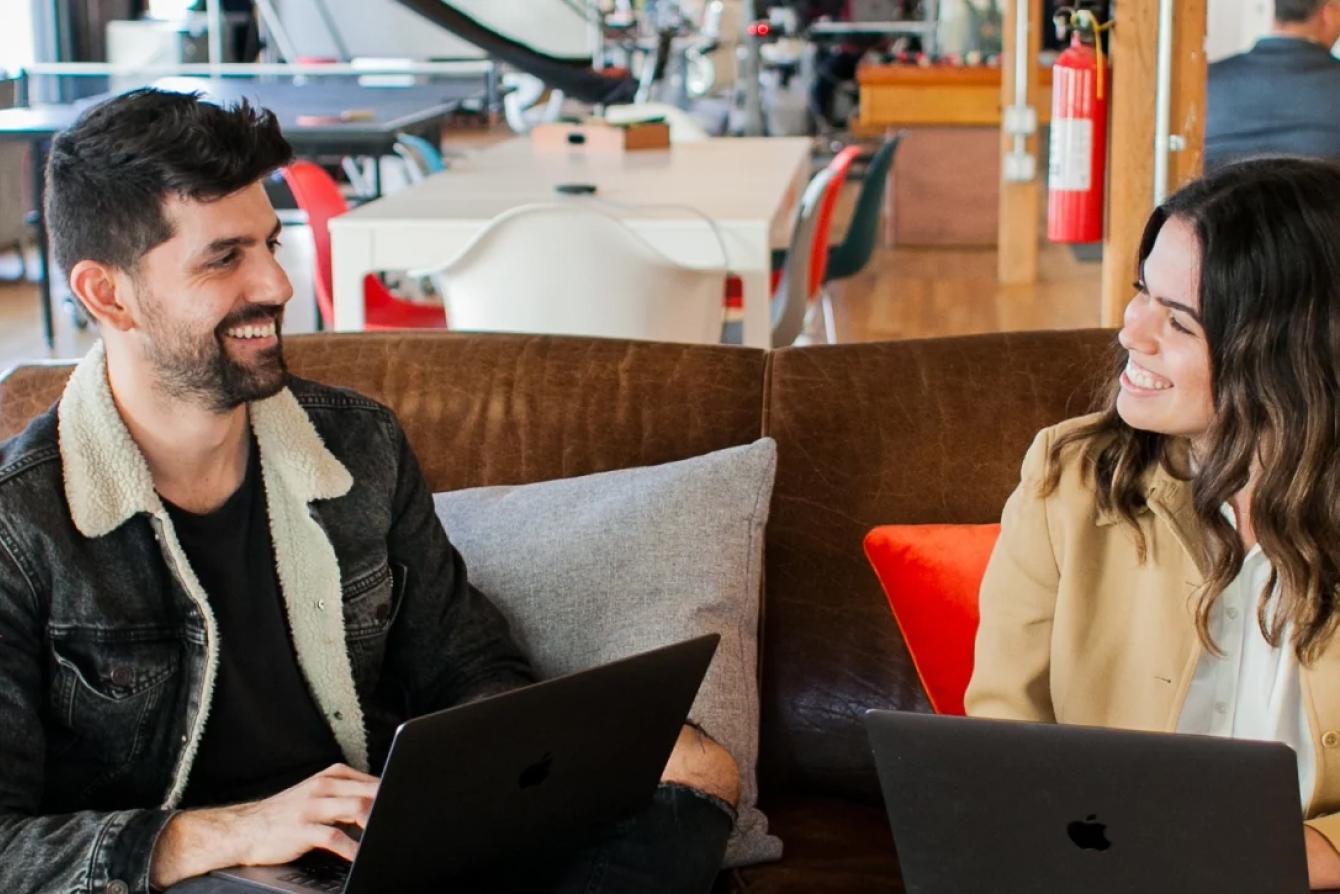 Two professionals having a casual meeting at a coffee shop, sitting on a brown couch with laptops while smiling at each other