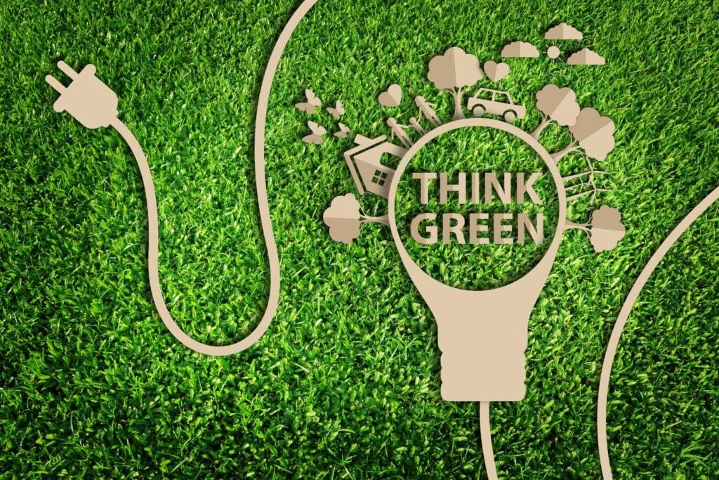 Paper cutout of lightbulb with 'Think Green' text and environmental icons on grass background with electrical plug