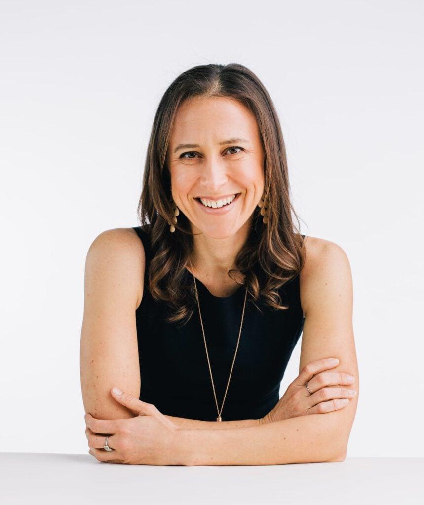 Professional woman in black sleeveless top smiling warmly at camera, wearing gold jewelry, against white background