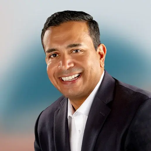 Professional headshot of a man in a dark suit jacket and white shirt smiling warmly against a blue gradient background