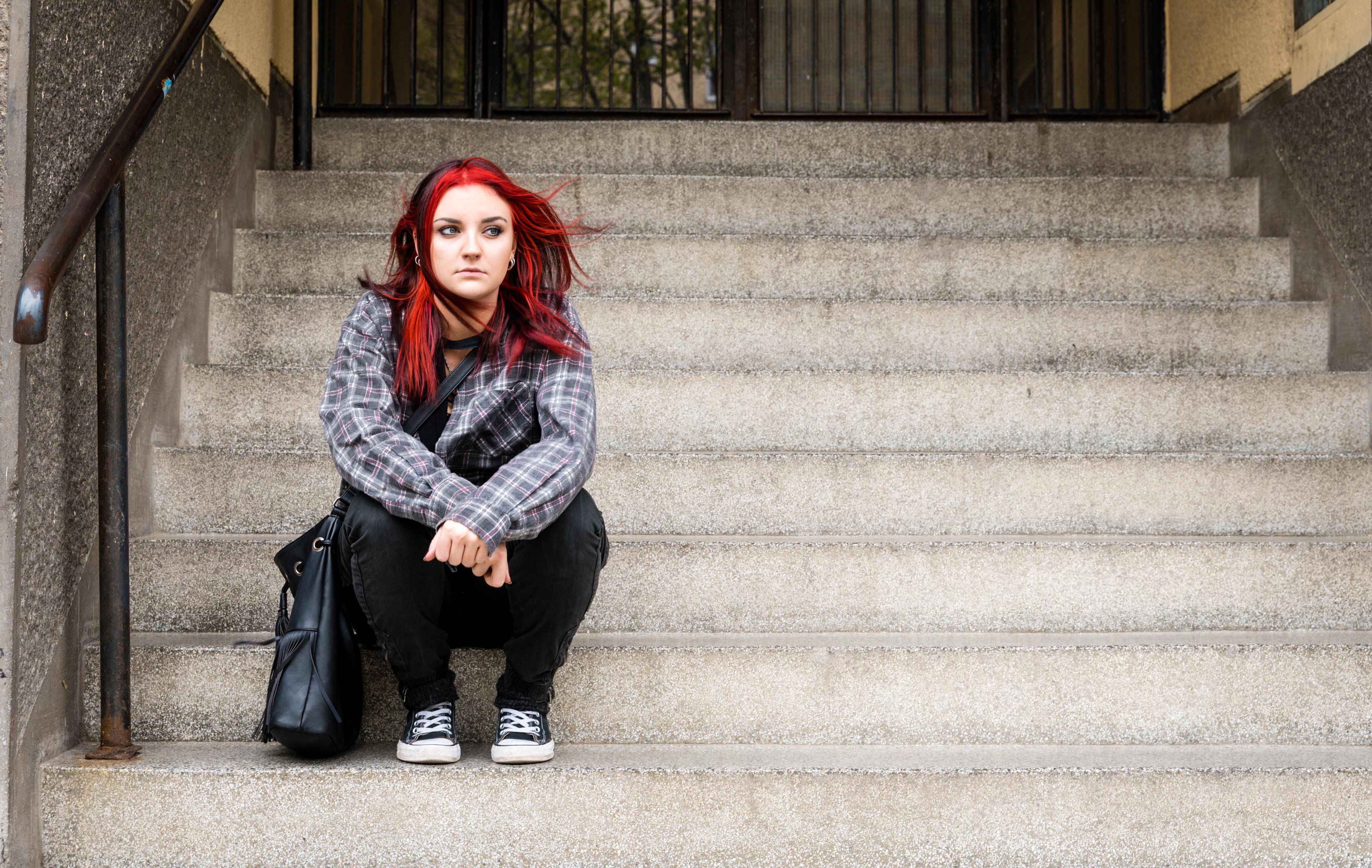 Young woman with bright red hair wearing plaid shirt and black pants, crouching on concrete steps with urban backdrop